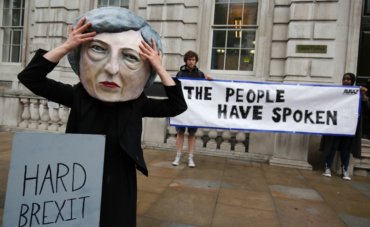 A demonstrator wears a mask depicting Britain's Prime Minister and leader of the Conservative Party Theresa May, poses with a mock gravestone bearing the words "Hard Brexit, RIP", during a protest photocall near the entrance 10 Downing Street in central London on June 9, 2017 as results from a snap general election show the Conservatives have lost their majority.