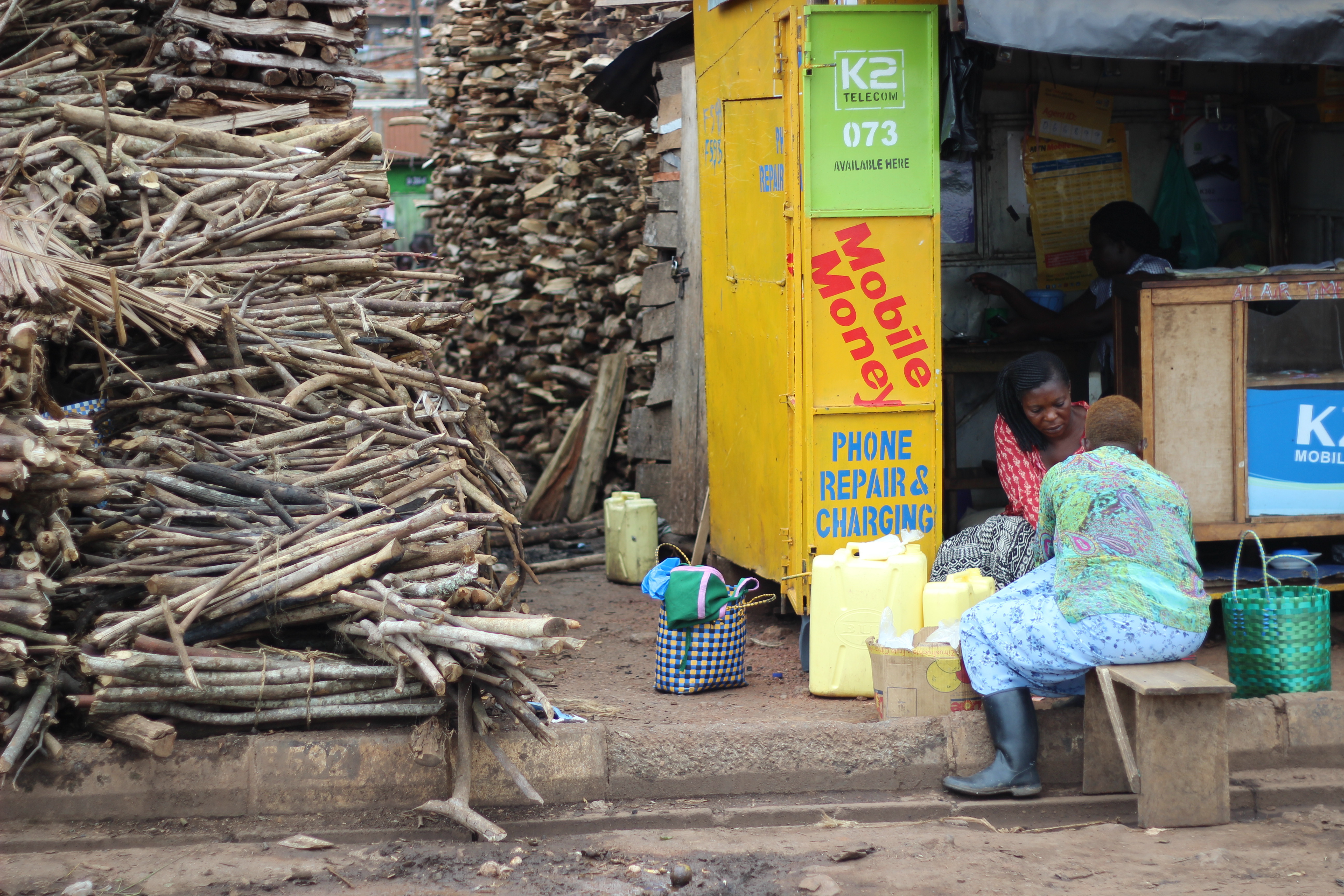 Women working at a market selling wood alongside operating a mobile phone repair shop in Kampala, Uganda