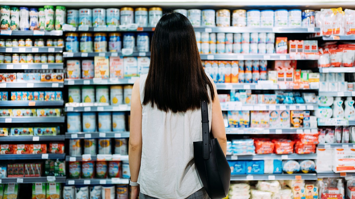 Rear view of young Asian mother groceries shopping for baby products in a supermarket. She is standing in front of the baby product aisle and have no idea which product to choose from