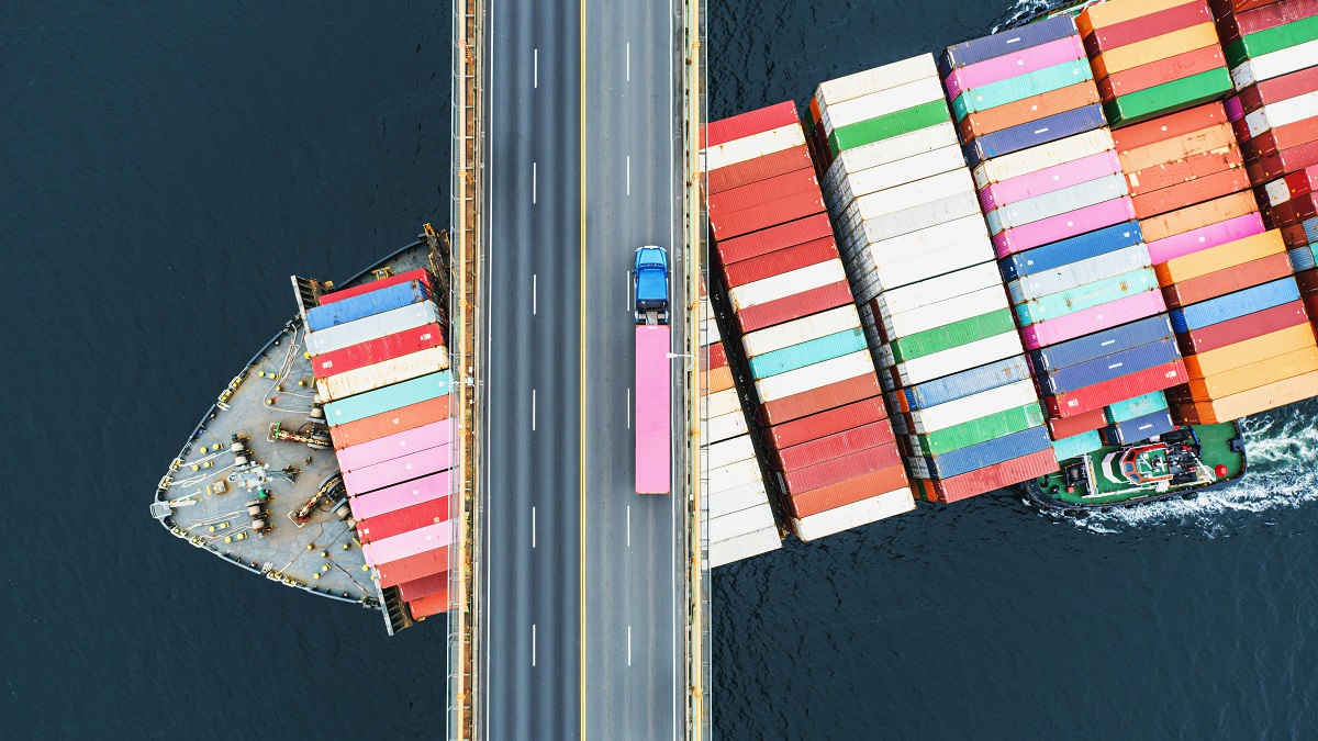 Aerial view of a container ship passing beneath a suspension bridge. Semi truck with pink cargo container crosses above.